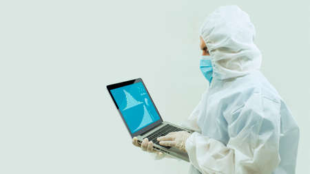 Doctor with white bioprotective suit and blue mask standing holding with his hands an open laptop...