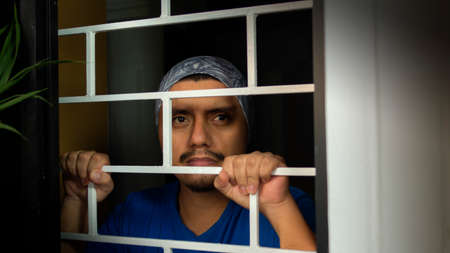 Close up view of  latin man with blue shirt locked in his house looking out through the window, leaning against security barsの写真素材