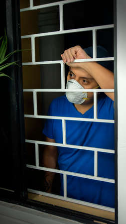 Latin man with white face mask and blue shirt inside his house looking out through the window, leaning against security barsの写真素材