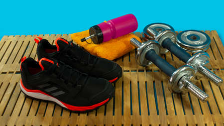 Gym set of pair of black sports shoes, dumbbells for exercise, yellow towel and red bottle with water on wooden floor with blue backgroundの写真素材