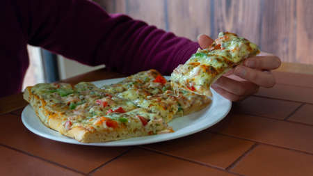 Woman's hand taking a rectangular piece of square pizza with ham, green pepper, tomato, olives and cheese from a white plate on a wooden tableの写真素材