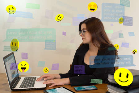 Beautiful Hispanic female office worker with glasses smiling happily sitting at her desk chatting her computer with speech bubbles and emojis floating around her against a white wallの写真素材