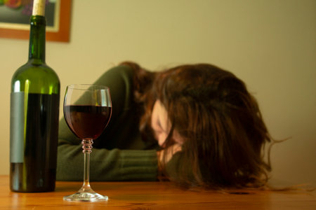 Close-up of a woman with a depressed expression in a green jacket sitting with her head on the table accompanied by a bottle and glass of red wineの写真素材