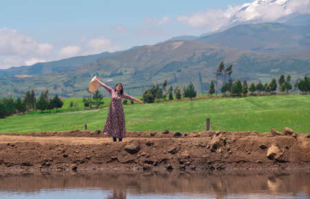 Beautiful Hispanic woman alone with flowered dress and hat raising her arms in happiness in the middle of a dirt road during a sunny day with blue skyの写真素材