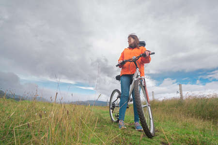 Young Hispanic woman seen from below wearing an orange jacket and blue pants resting on her bicycle in the middle of a rural field during a cloudy dayの写真素材