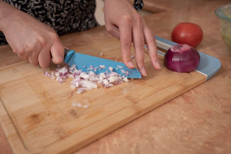 Close-up of a woman's hands chopping onions with a blue knife on a wooden board at the kitchen...