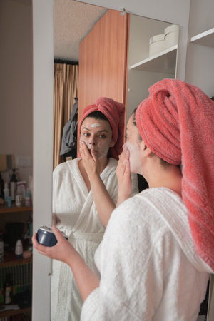 Beautiful Hispanic woman in a white bathrobe and red towel in her hair applying makeup with her hand on her face in front of the mirror on the closet door in her bedroomの写真素材