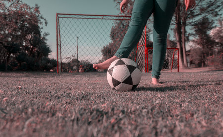 Close up on legs of woman in blue pants, barefoot in middle of park playing with a soccer ball with a soccer goal with nets and trees in background during the morning of sunny day. Retro styled imageの写真素材