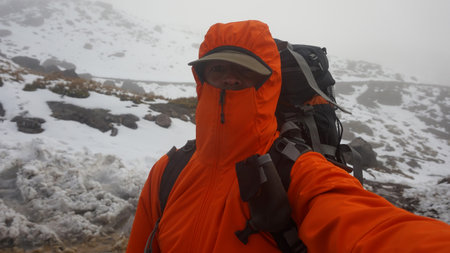 Young climber man with backpack, red coat and cap taking a selfie while climbing a snowy mountain on...