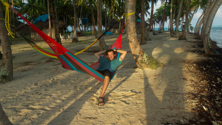 Young tourist man lying in a hammock surrounded by palm trees by the sea during a sunny dayの写真素材