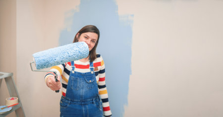 Close-up of roller with blue paint in the hand of a standing woman dressed in overalls and striped blouse, seen from the front, against background of half-painted white wall with strokes of blue paintの写真素材