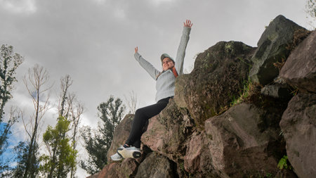 Beautiful young Latin American woman sitting on stones at the top of a mountain with her arms raised...