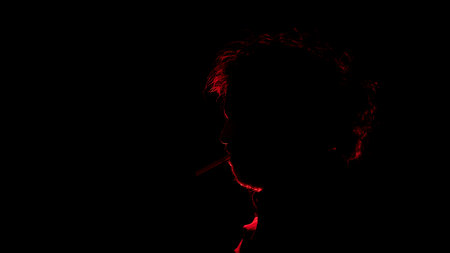 Portrait of Adult man with a cigarette in his mouth seen in profile in silhouette with red light illuminating his profile against black backgroundの写真素材