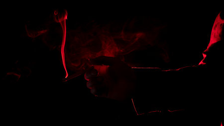 Close-up of a man's hand with a lit cigarette, blowing smoke, seen from profile in silhouette with red light illuminating his profile against black backgroundの写真素材