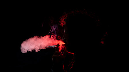Portrait of Adult man with a cigarette in his hand, blowing smoke out of his mouth seen in profile in silhouette with red light illuminating his profile against black backgroundの写真素材