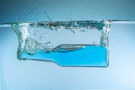 Clear glass bottle filled with blue liquid submerged in water causing a splash against a blue backgroundの写真素材