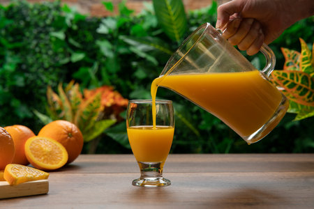 Man's hand with orange juice jug filling a glass cup on a wooden table against a background of green plantsの写真素材