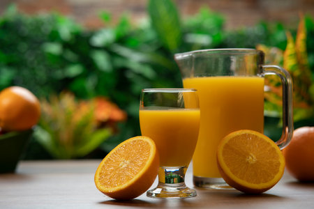 Half an orange resting on a glass of juice and a jug of orange juice on a wooden table against a background of green plantsの写真素材