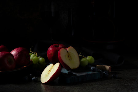 Red apples, green grapes, and blueberries on a chopping board on a black ceramic table with wine bottles in the dark backgroundの写真素材