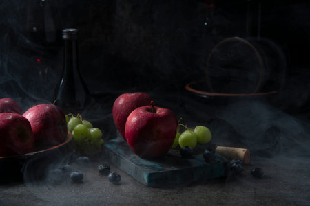 Red apples, green grapes, and blueberries on a chopping board with fog around it on a black ceramic table with a bottle of wine in the dark backgroundの写真素材