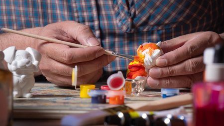 Close-up of a man's hands in a blue shirt holding a fine brush with orange paint, painting a small...