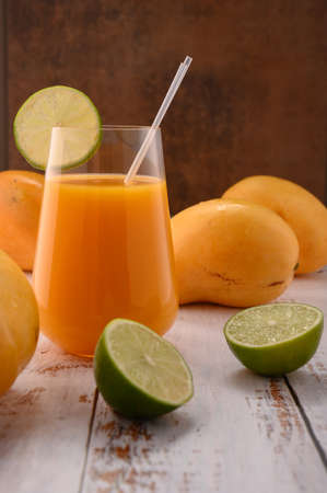 Photo shows glass transparent with natural fresh mandarine juice on vintage textured. In background, exotic yellow mango fruits can be seen. In the foreground there is slices of citrus fruit - lime.の写真素材