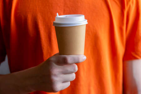 A paper cup of takeaway coffee in the hand of a young man in a bright orange T-shirtの写真素材