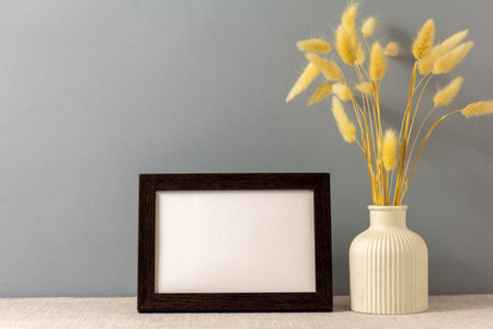 The layout of an empty photo frame on the table. A vase with dry grass. A minimalistic workspace. A...