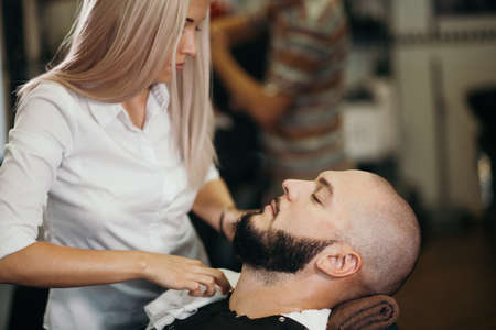 Portrait of handsome smiling young bearded caucasian man getting trendy haircut in modern barbershop. Attractive barber girl working, serving client, doing haircut using shaver.の写真素材