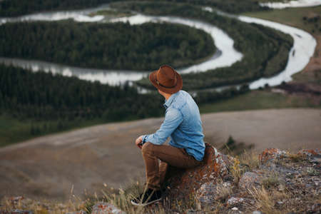 picture of a young fashion man posing outdoor with a bag at his feet while sitting on a rock and looking away from the camera, with a beautiful landscape behind himの写真素材