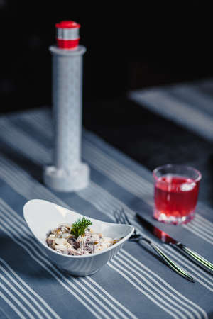 Table in the restaurant. On the table is a salad in a white plate, a knife, a fork, a glass with red juice and a small lighthouse. On the table is a blue tablecloth in strips. Black background.の写真素材