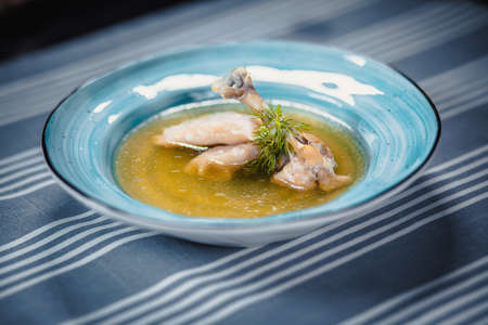Soup in blue plate with chicken broth, chicken's leg, dumplings and greenery. Plate on the table with a blue tablecloth in white strips. Clouse up.の写真素材