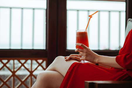 Close up of woman in red dress with red cocktail with orange straw in hands. Beautiful girl in red dress drinking red cocktail from a glass on an open terrace of an expensive fashionable restaurant.の写真素材