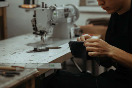 Close up of leather craftsman working with natural leather and tools. Handmade master at work in local workshop. Leather wallet stitch.の写真素材