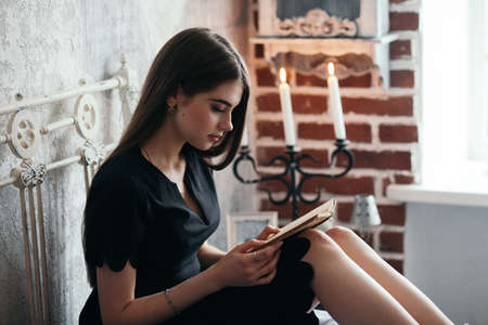 A girl is reading a book on a bed in a romantic black dress.