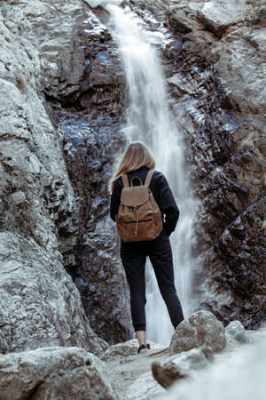 Girl with backpack standing on rocks and looking at waterfall