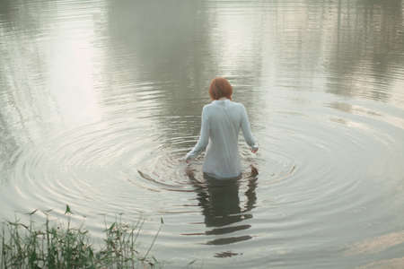 Beautiful red-haired woman standing in lake in misty forestの写真素材