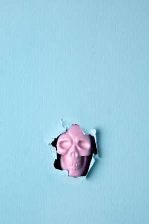 Halloween pink skull breaking through pastel blue wall. Minimal holiday celebration funny concept. Bright autumn season background.の写真素材