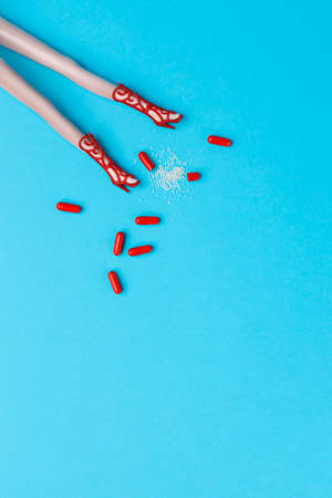 Creative layout made with doll legs and red pills on blue background. Minimal medical or tablet abuse concept. Flat lay.の写真素材