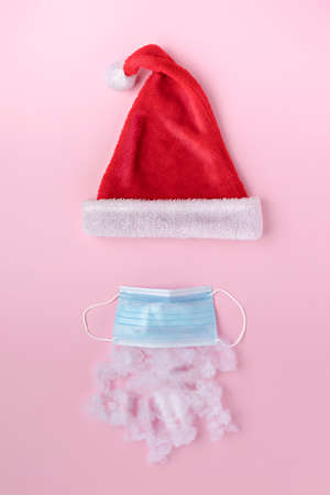 Creative Christmas layout with Santa's face made of surgical mask, beard and hat on pink background. New Year, winter, isolation, coronavirus and stay at home quarantine concept. Flat lay, top view.の写真素材