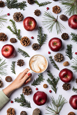 Creative Christmas layout made with hand holding beverage, fir, apples, branches and pine cone on...