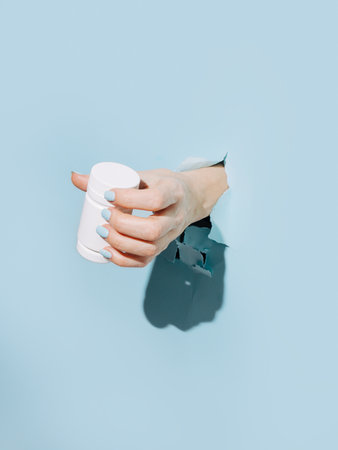 Female hand holding pill bottle breaking through pastel blue wall. Mock up or template for empty bottle. Trend summer shadow visual concept.の写真素材