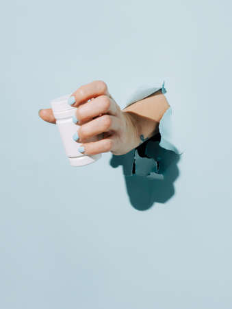 Female hand holding pill bottle breaking through pastel blue wall. Mock up or template for empty...