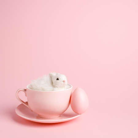 Easter bunny rabbit in a teacup with painted egg on pastel pink background. Creative Easter holiday concept with copy space. Minimal spring holiday greeting card.の写真素材