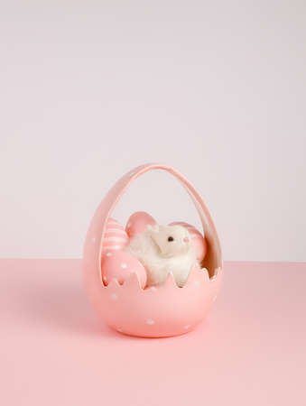 Fluffy white bunny in egg shaped pink basket with painted eggs on two tone pastel bright background. Minimal Easter concept.の写真素材