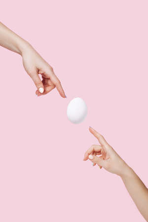 Two hands touching flying egg on pastel pink background. Creation of new life. Minimal Easter art concept.の写真素材