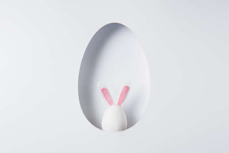 Easter border template with cut in egg shape and small white egg wearing bunny ears on bright background. Minimal Easter holiday greeting card or banner with frame for copy space. Flat lay.の写真素材