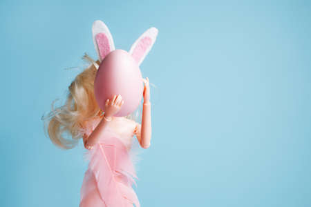 Doll with bunny ears and pink feather dress holding egg against pastel blue background. Creative Easter or fashion concept.の写真素材