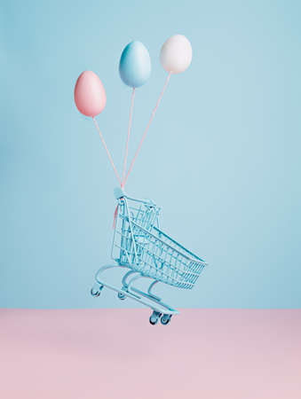 Painted egg helium balloons and shopping cart floating against two tone pink and blue background. Creative Easter shopping concept.の写真素材
