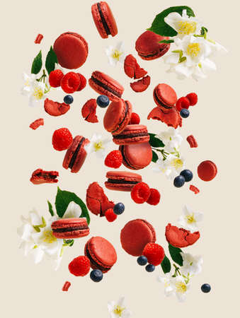 Red macaroon cakes falling with raspberries, flowers, and blueberries on a bright beige background. Creative food concept. Crispy and creamy cakes with summer fruits.の写真素材